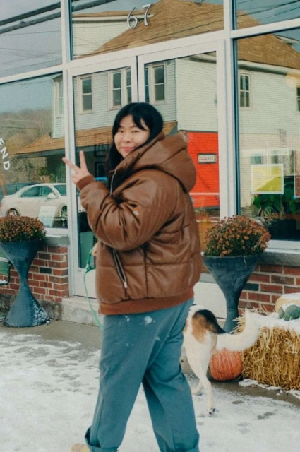 Asian woman in brown puffer jacket flashes peace sign in snowy urban setting with dog, plants, buildings reflected in window.