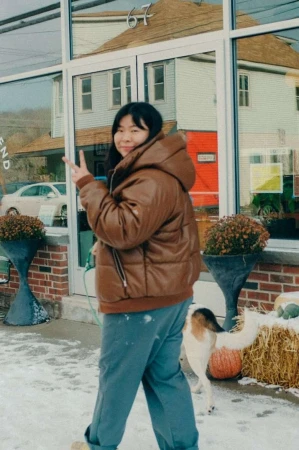 Asian woman in brown puffer jacket flashes peace sign in snowy urban setting with dog, plants, buildings reflected in window.