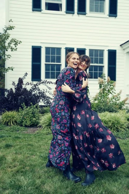 Two women embrace outdoors in floral dresses. They're standing on green grass in front of a white house with dark shutters and green plants.