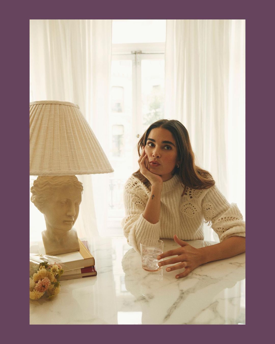 Portrait of a woman in a cream sweater sitting at a table with a bust, books, a lamp, and a glass in a brightly lit room.