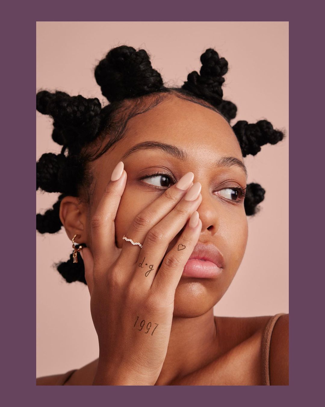 Portrait of a young Black woman with distinctive Bantu knots hairstyle, featuring a tan top, delicate jewelry, and hand tattoos.