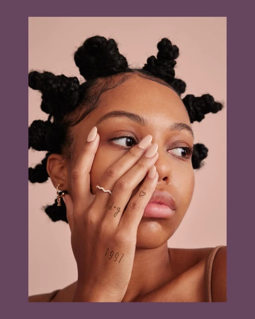 Portrait of a young Black woman with distinctive Bantu knots hairstyle, featuring a tan top, delicate jewelry, and hand tattoos.
