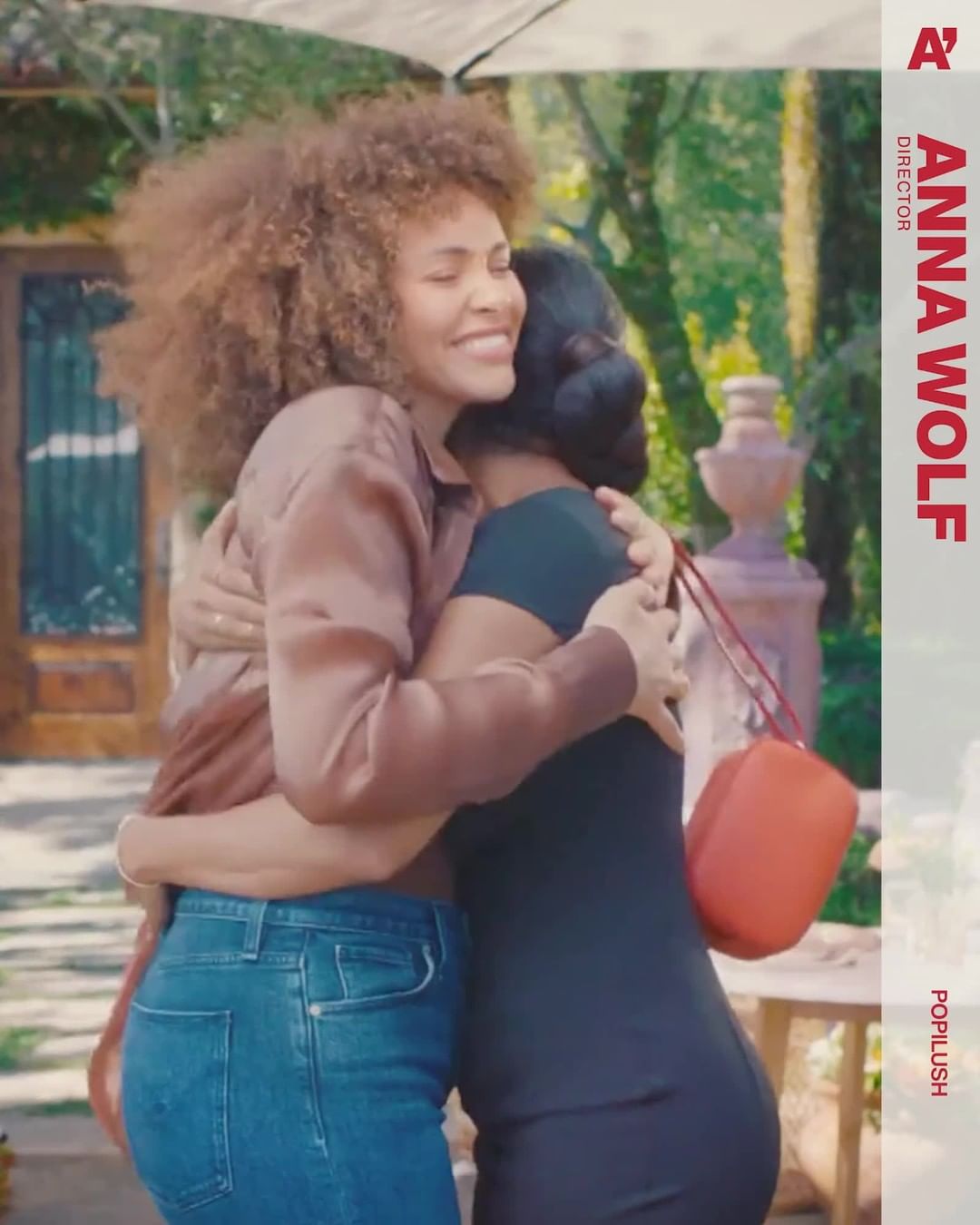 Two women embrace outdoors; one with afro and jeans, the other with a black dress and purse, set against a backdrop of lush greenery.