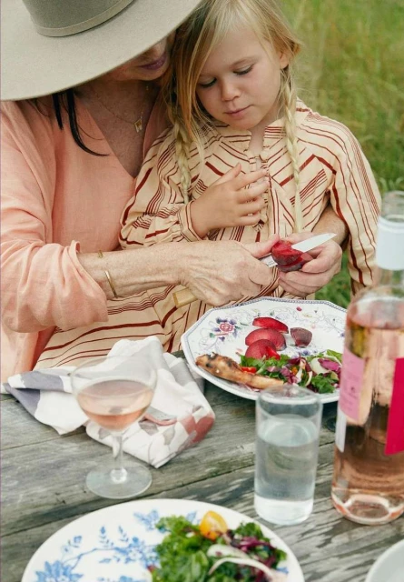 Image of a woman and child outdoors at a picnic table, the woman cuts a plum, salads and drinks sit on a weathered wood table.