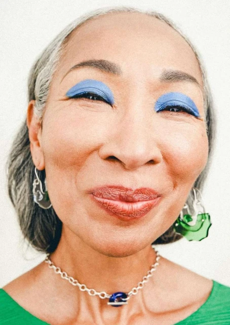 Close-up portrait of a smiling, mature Asian woman with blue eyeshadow, silver jewelry, and green top against a white background.