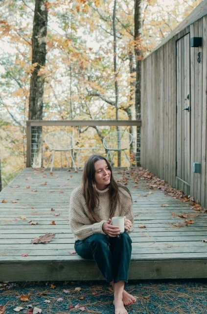 Woman sits on wooden steps holding mug in autumnal forest. She wears a cream-colored sweater and dark pants in a natural outdoor setting.