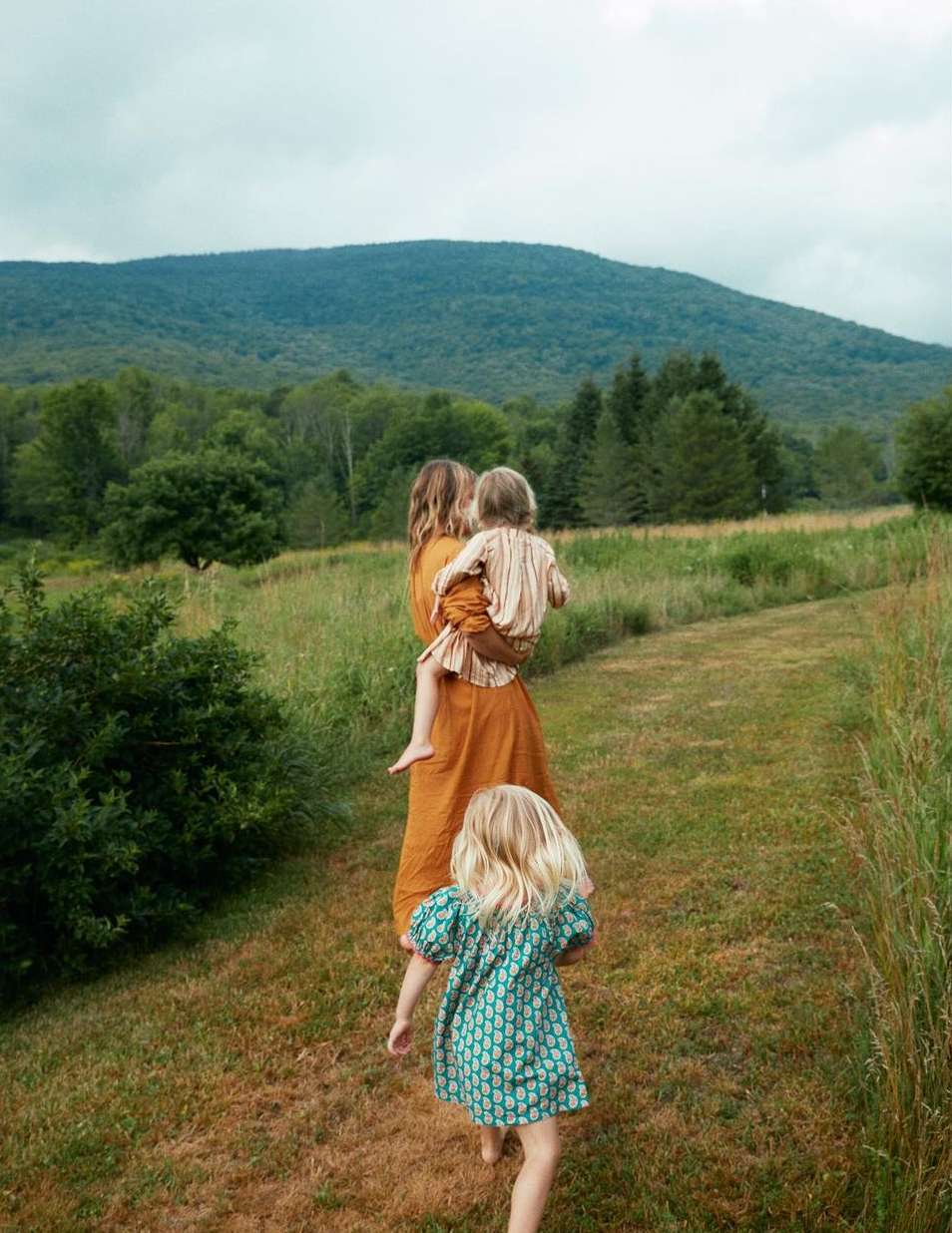 Mother and two young daughters walking in a grassy field with a mountain backdrop on a cloudy day, enjoying nature and family time.