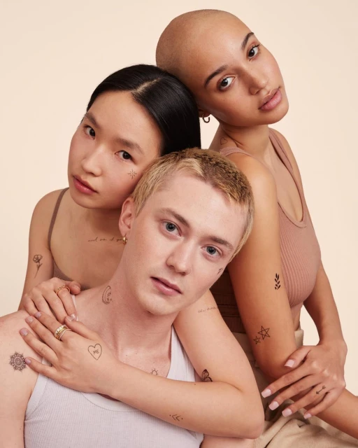 Three diverse young adults pose closely together, showcasing temporary tattoos against a soft beige backdrop in a studio setting.
