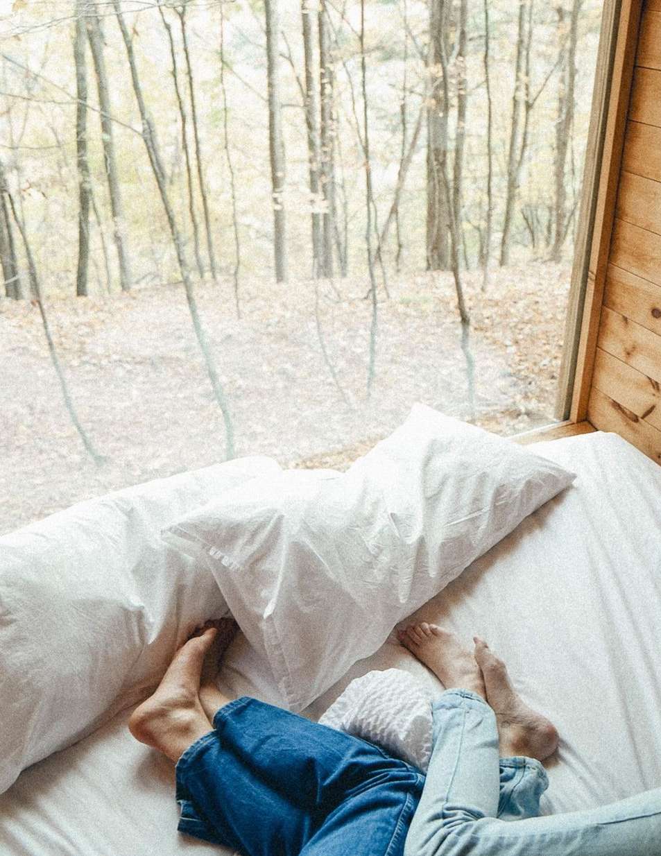 View from a cabin interior featuring a bed with two people's feet visible, looking out through a large window at a forest scene.