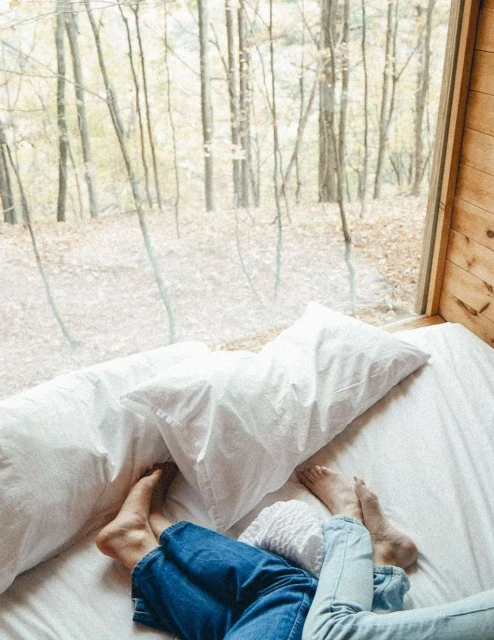 View from a cabin interior featuring a bed with two people's feet visible, looking out through a large window at a forest scene.