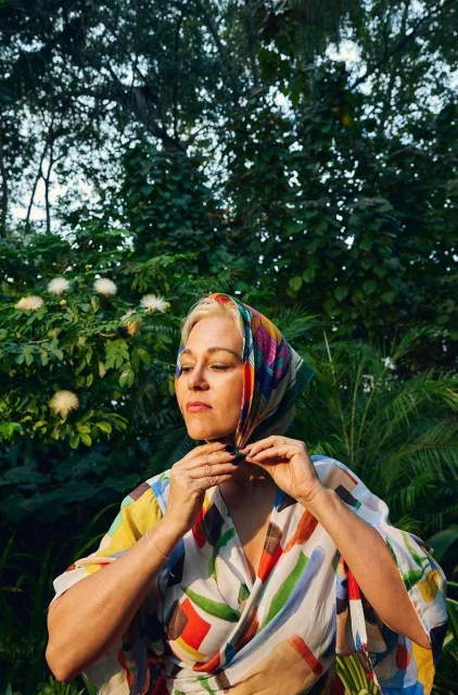 Adult woman in a colorful headscarf and printed wrap standing outdoors amid green trees and foliage in natural lighting.