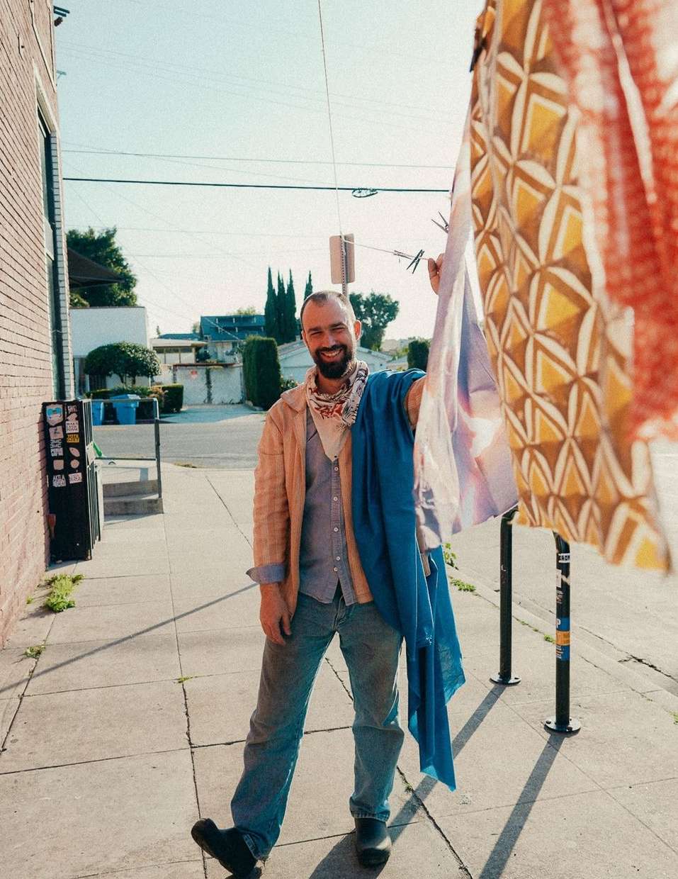 Outdoor portrait of a smiling man holding patterned fabrics. He is standing on a sidewalk with buildings visible in the background.