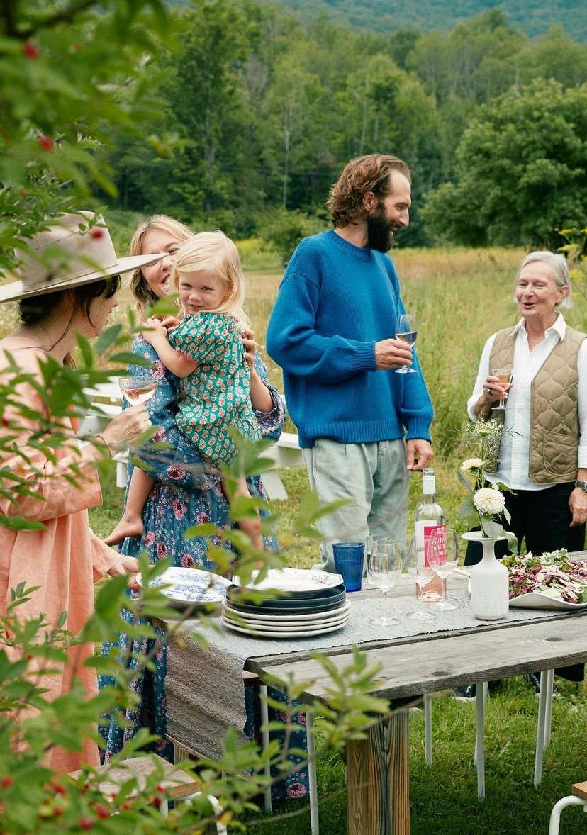 Group of family and friends gathered around a wooden outdoor dining table in a green meadow, photographed by Anna Wolf.