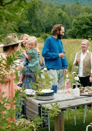 Group of family and friends gathered around a wooden outdoor dining table in a green meadow, photographed by Anna Wolf.