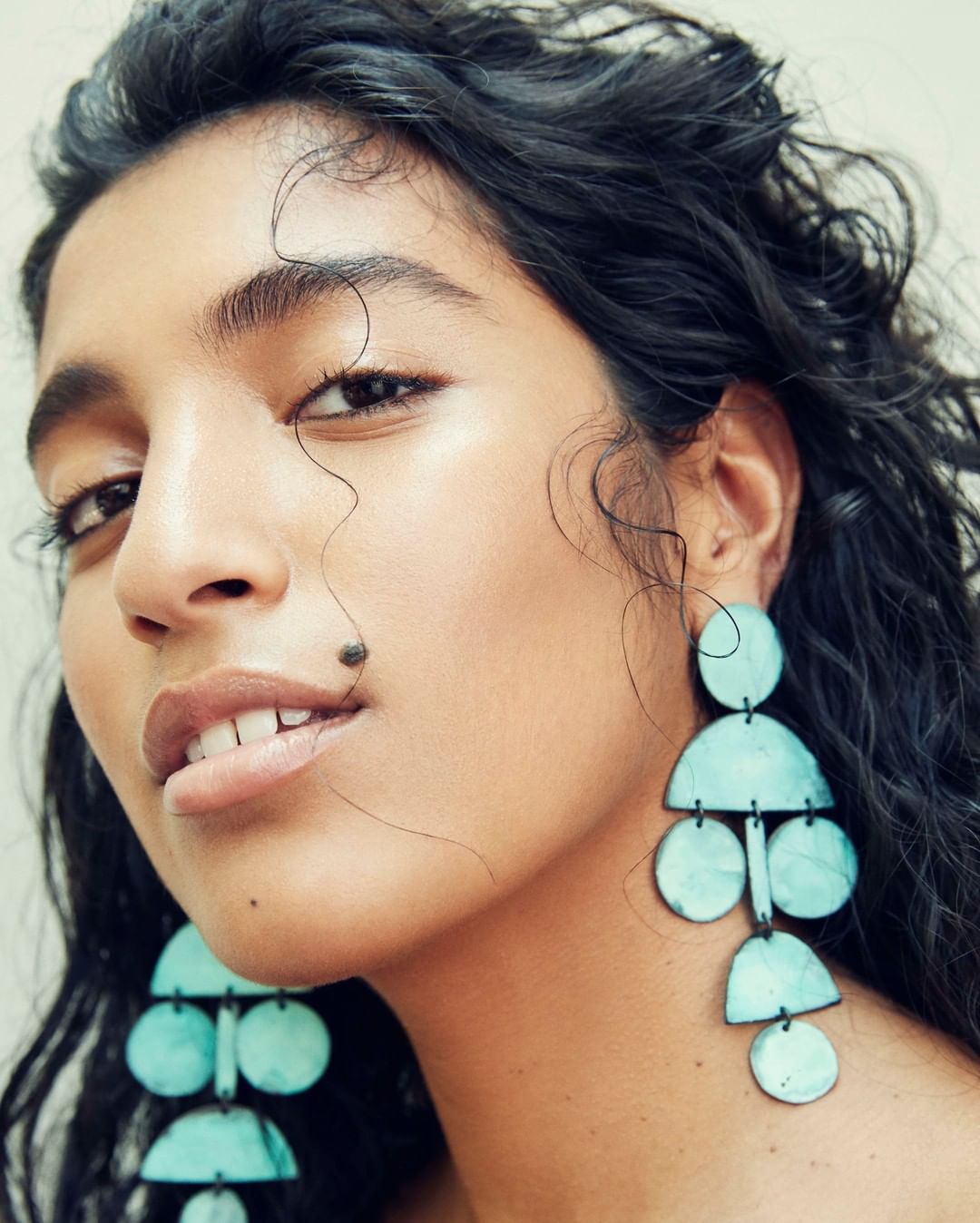 Close-up of a woman with dark curly hair and large, turquoise, geometric earrings. Beauty shot with focus on her face and jewelry.