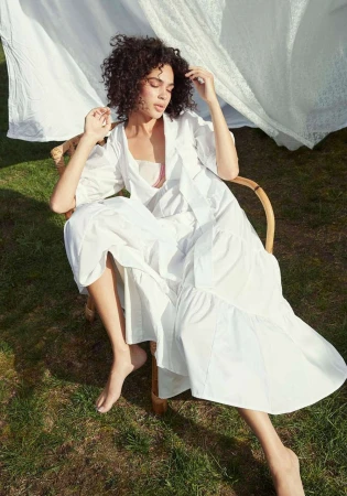 Woman with curly dark hair wearing a long white dress seated in a wicker chair outdoors with white fabric draped behind her
