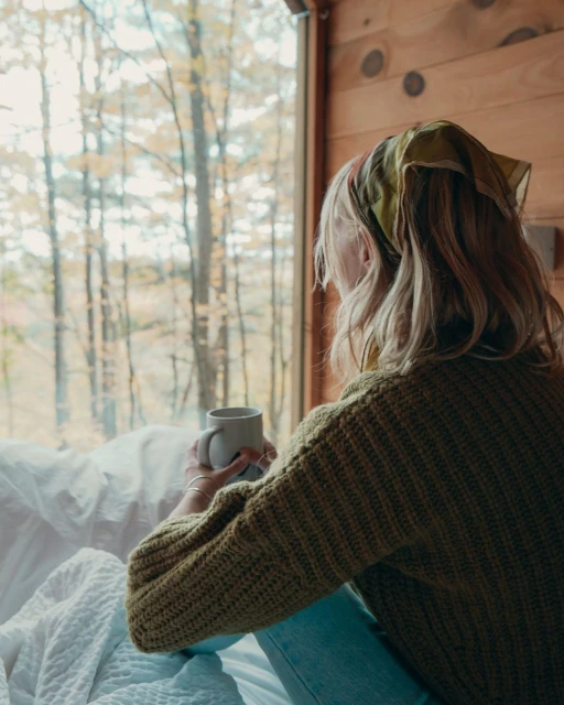 Blonde woman in sweater and headscarf looks out window at forest with coffee mug in hands, sitting in white bed inside cabin.