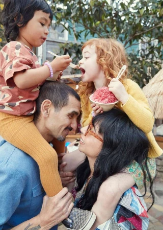 A family of four poses outdoors; the two children sit on their parents' shoulders, enjoying a snack, set against a blurred background.