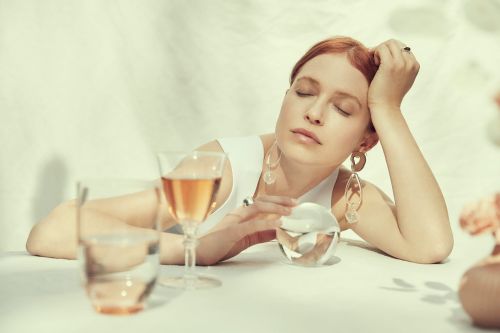Red-haired woman with eyes closed sits at a table with drinks and jewelry, illuminated by natural light and soft shadows.
