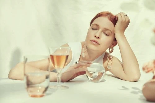 Red-haired woman with eyes closed sits at a table with drinks and jewelry, illuminated by natural light and soft shadows.