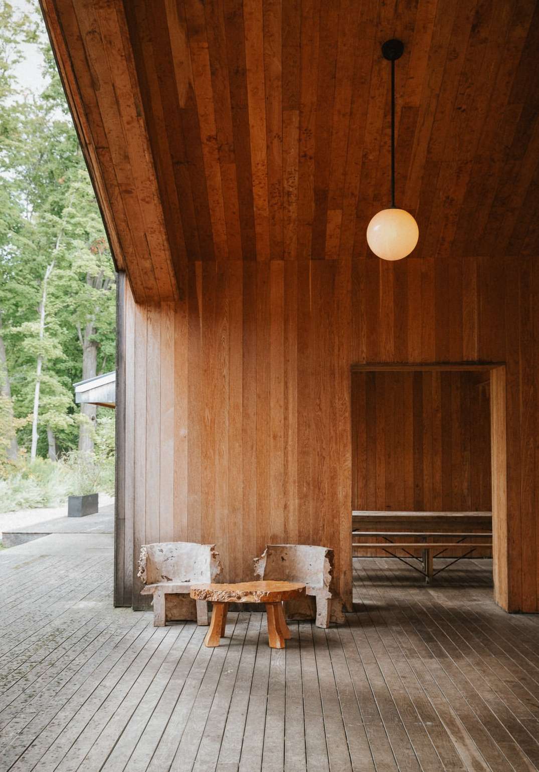Interior view of a wooden building with rustic chairs, a wooden table, and a pendant light, set against a green forest backdrop.