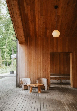 Interior view of a wooden building with rustic chairs, a wooden table, and a pendant light, set against a green forest backdrop.