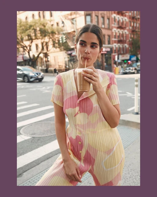 A woman wearing a yellow floral jumpsuit drinks from a cup with a straw in a city setting with buildings and a crosswalk.