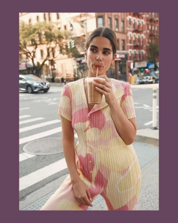 A woman wearing a yellow floral jumpsuit drinks from a cup with a straw in a city setting with buildings and a crosswalk.