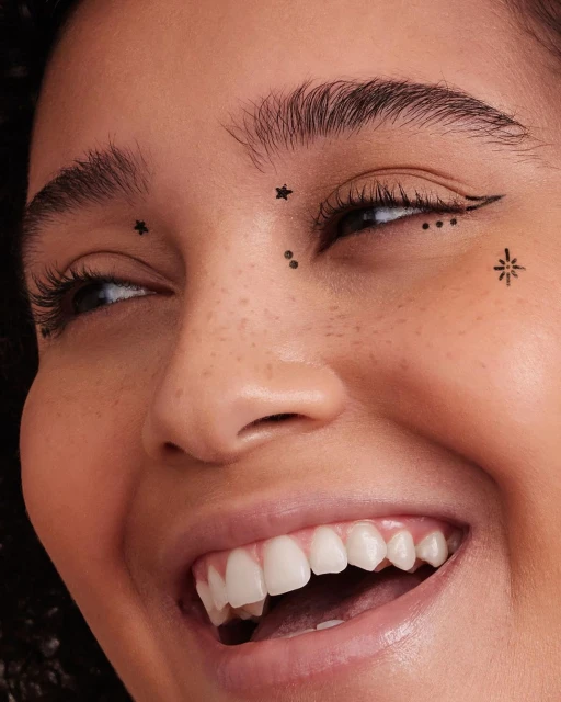 Close-up of an adult woman with eye makeup, including graphic eyeliner and star and dot face art, smiling with white teeth.