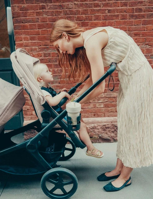 Mother and toddler interact near stroller on brick sidewalk. Woman with long red hair wears a dress and flats; toddler wears sandals.