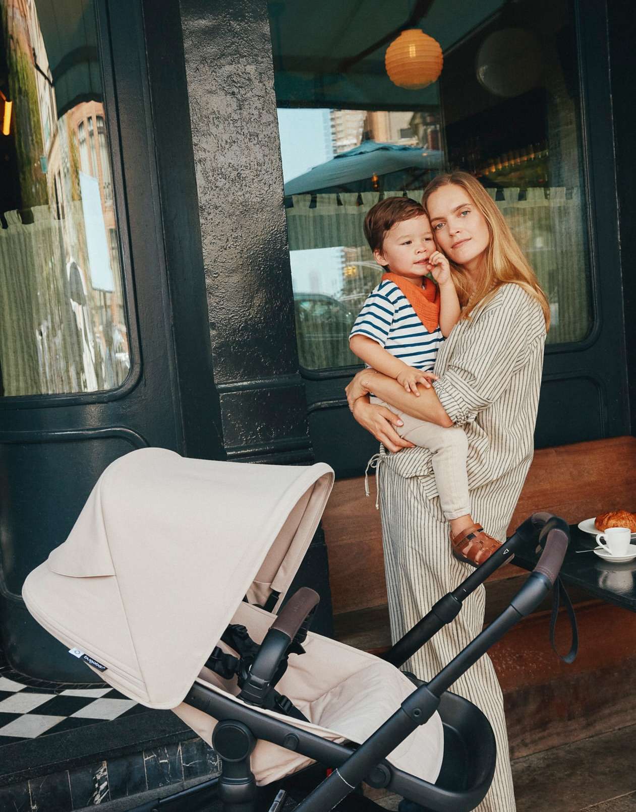 A young woman holds a toddler next to a black stroller in front of a storefront with a table holding a croissant and coffee.