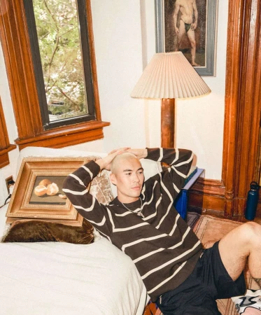 Man with shaved blonde hair seated on a white couch wearing a brown striped sweater, photographed by Anna Wolf