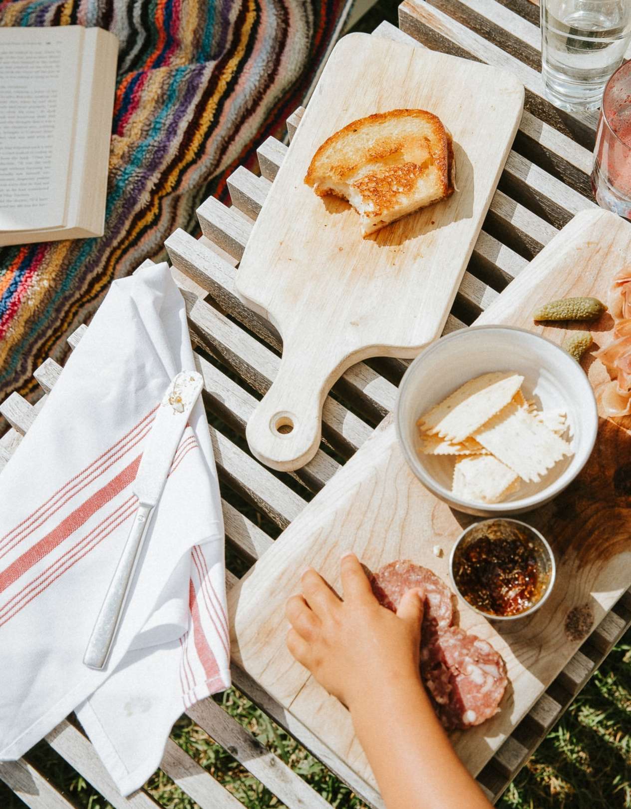 Overhead shot of a young child reaching for salami on a wooden board with toast, crackers, and relish, all set on a wooden table.