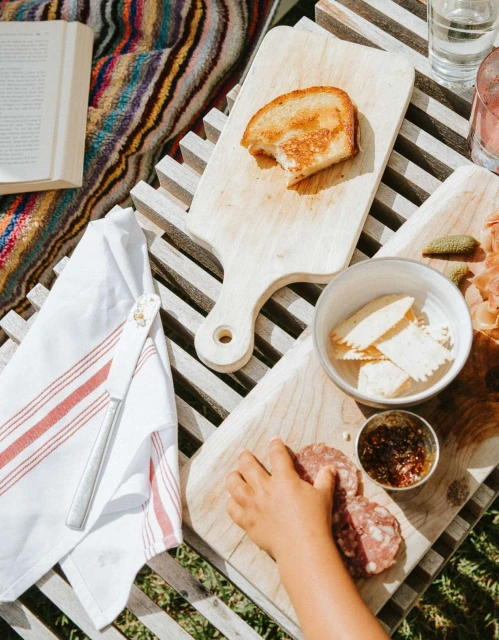 Overhead shot of a young child reaching for salami on a wooden board with toast, crackers, and relish, all set on a wooden table.
