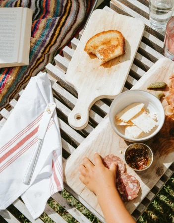 Overhead shot of a young child reaching for salami on a wooden board with toast, crackers, and relish, all set on a wooden table.