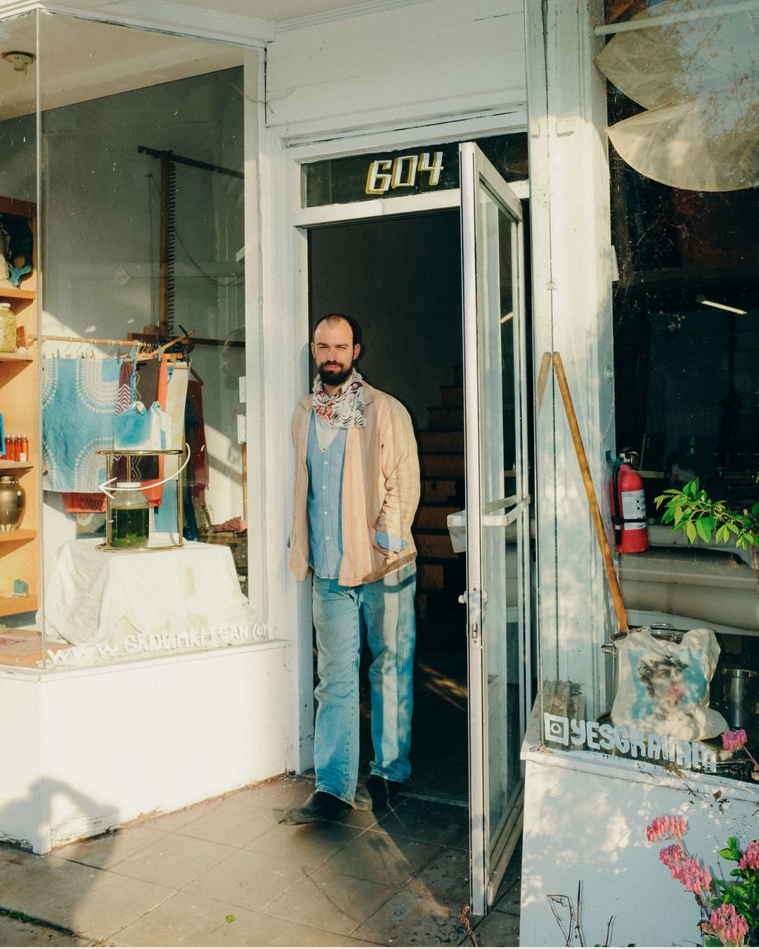 Adult male in light blue shirt and jeans stands in open doorway of a building with street-facing window display and building number 604.