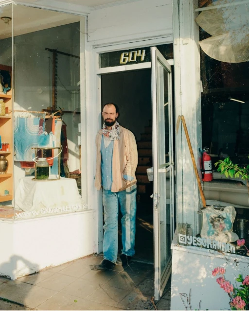Adult male in light blue shirt and jeans stands in open doorway of a building with street-facing window display and building number 604.