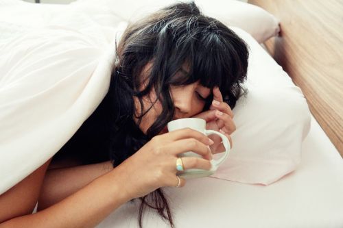 Woman in bed with dark hair drinking from a white ceramic mug on white pillows