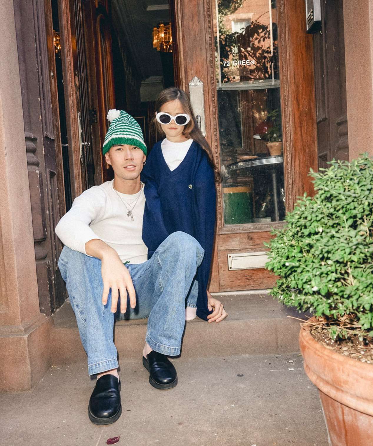 Young adult and child pose on brownstone stoop in front of a dark wooden door, with natural light and casual street style.