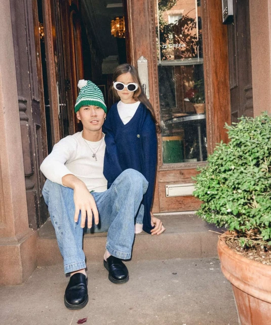 Young adult and child pose on brownstone stoop in front of a dark wooden door, with natural light and casual street style.