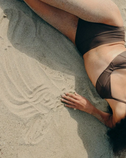 Woman in a brown bikini lies on a sandy beach with visible handprints and sunny lighting, creating a peaceful and relaxed beach scene.