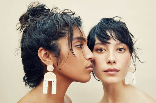Two women pose with neutral makeup and statement earrings in a studio shot with warm lighting. Beauty and fashion concept.