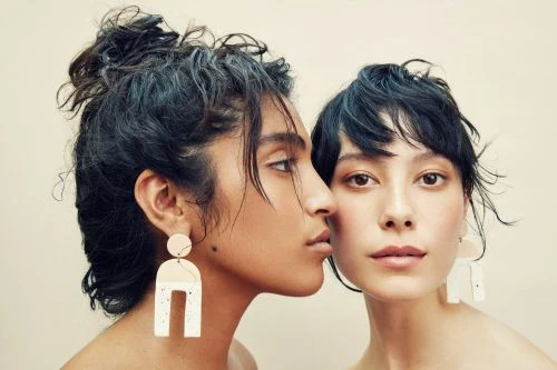 Two women pose with neutral makeup and statement earrings in a studio shot with warm lighting. Beauty and fashion concept.