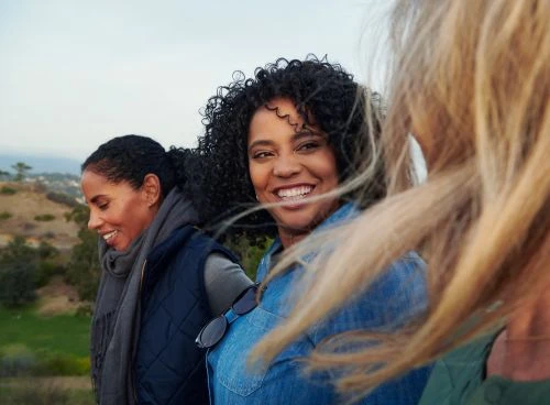 Three women smiling outdoors with one African-American woman in focus, wearing a denim shirt and sunglasses, nature background.