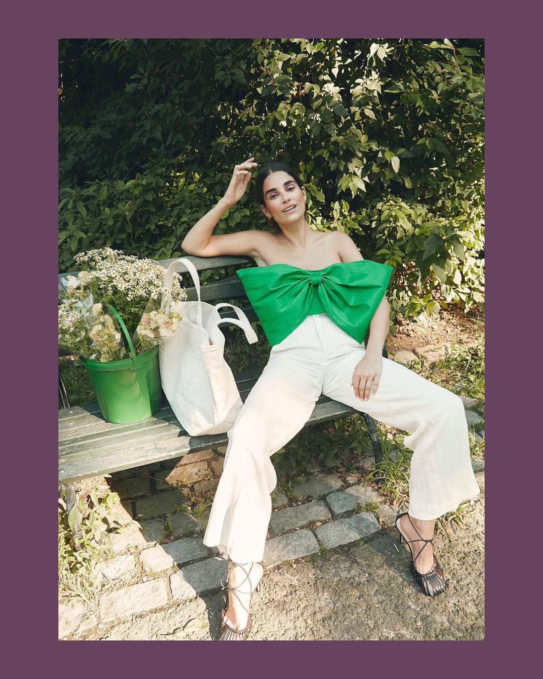 Brunette woman in a green bow top and white pants sits on a park bench with a bucket of flowers and tote bag in an outdoor setting.