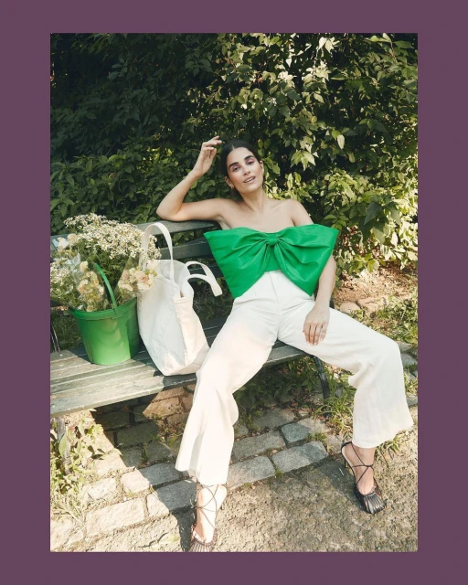 Brunette woman in a green bow top and white pants sits on a park bench with a bucket of flowers and tote bag in an outdoor setting.