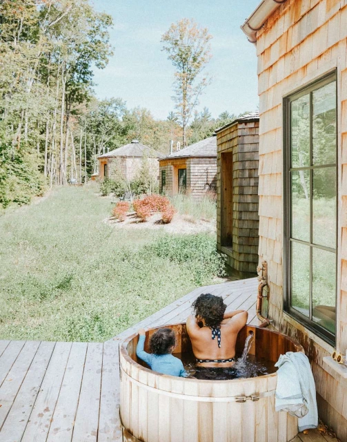 Adult and child relax in an outdoor cedar hot tub, surrounded by wooden buildings and a green forest, on a sunny day.
