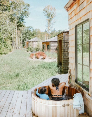 Adult and child relax in an outdoor cedar hot tub, surrounded by wooden buildings and a green forest, on a sunny day.