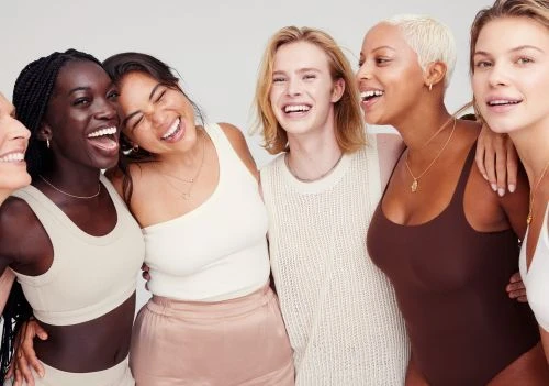Group of six diverse women laughing, wearing casual tops and minimalist necklaces, posing together against a clean, neutral background.