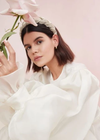 Woman in white blouse wearing a braided headband holding a pink rose against a pale pink background by Anna Wolf.